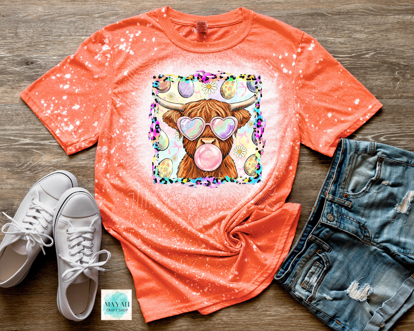 Orange t-shirt with a colorful cow graphic, white sneakers, and denim shorts on a wooden surface.