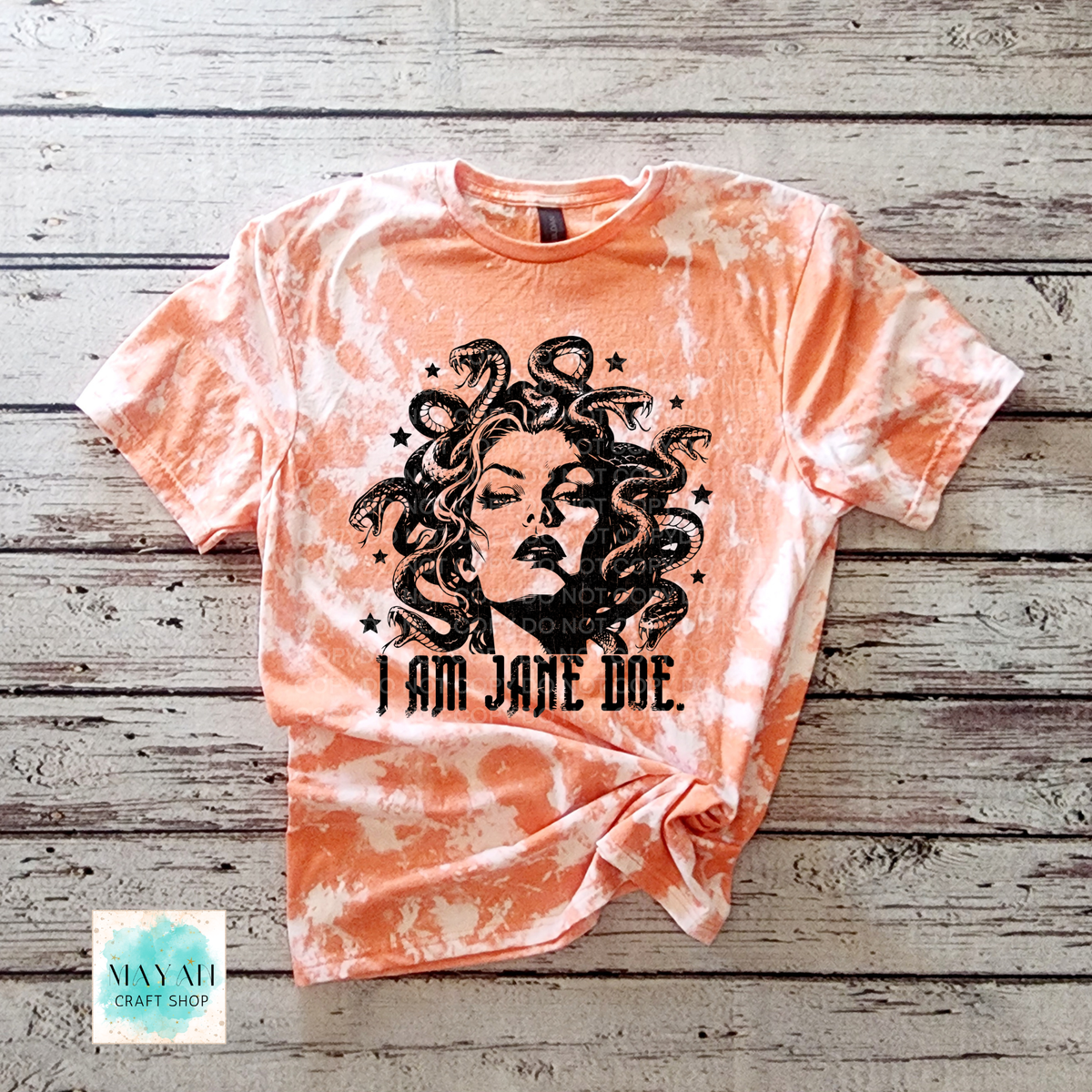 A heather orange bleached graphic t-shirt featuring a black illustration of Medusa and the text 'I AM JANE DOE.' laid flat on a rustic gray wood plank background.