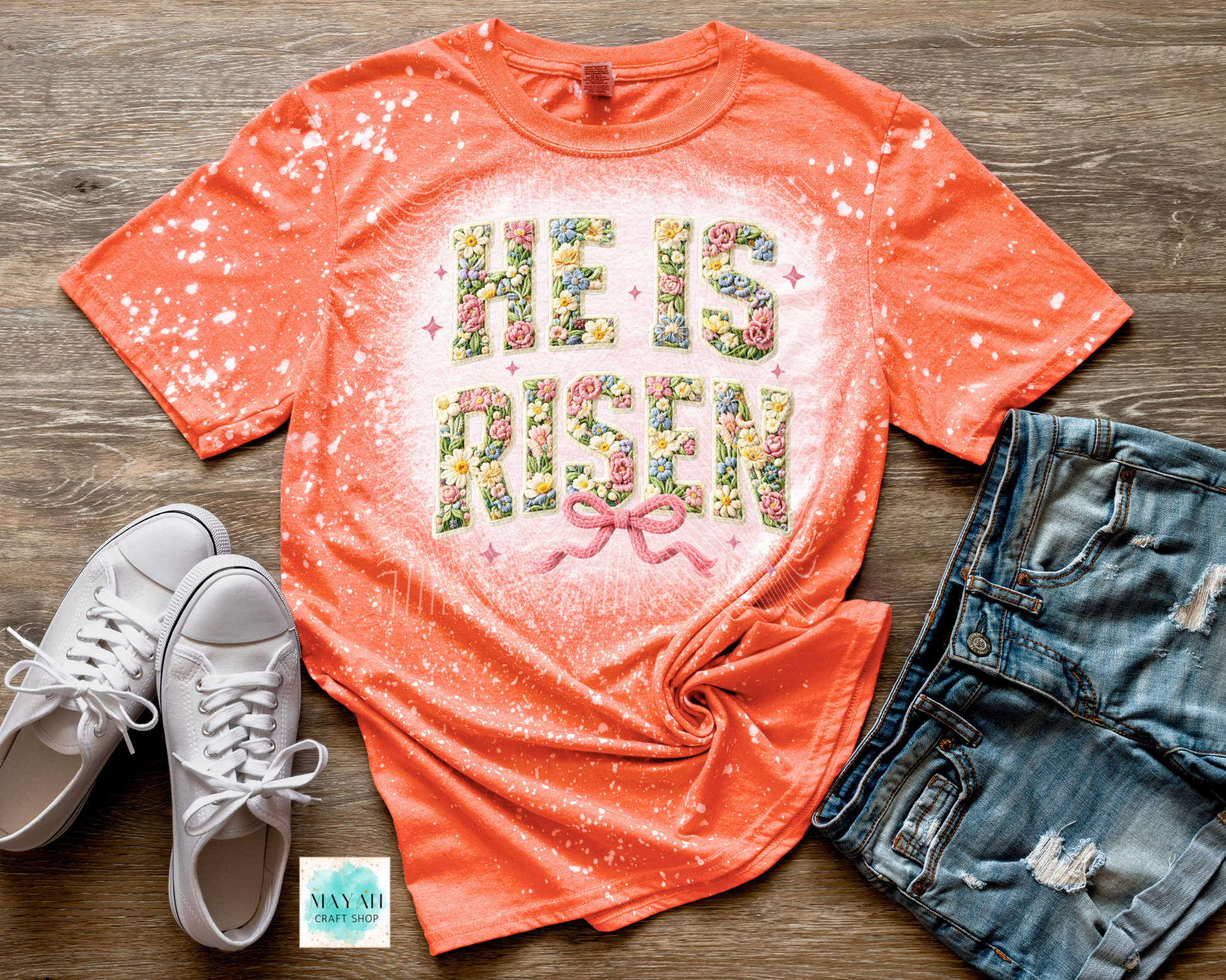 Coral t-shirt with 'He is Risen' text, white sneakers, and denim shorts on a wooden surface.