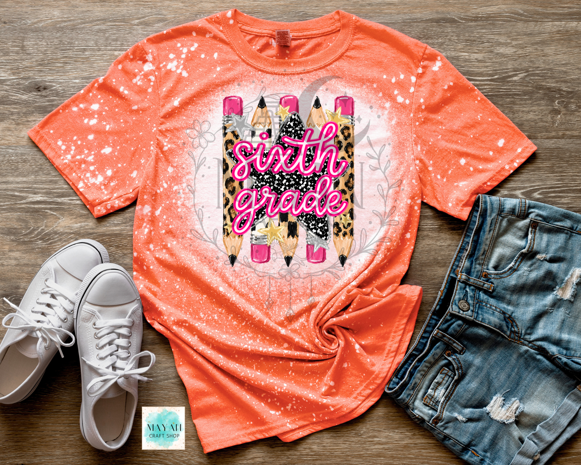 An orange short-sleeve graphic T-shirt laid flat on a wood surface, styled with white sneakers, distressed denim shorts, and a small card. The shirt has a splatter/glitter finish and a central graphic of pencils with the words 'sixth grade' in pink script over leopard-print and glittery pencil bodies.