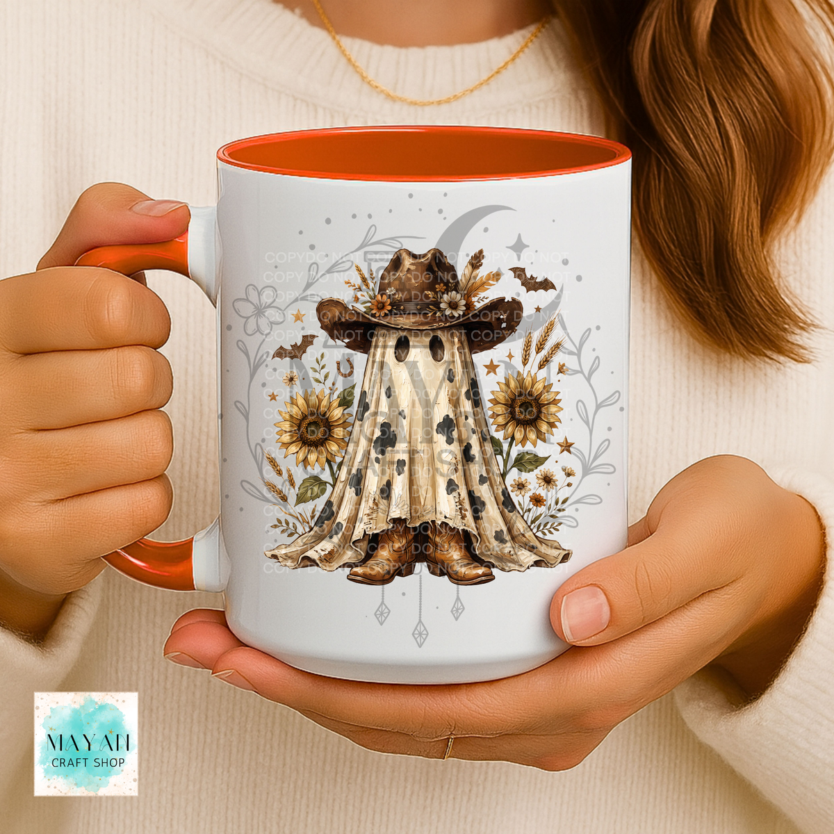 A person holds a white ceramic mug with a bright orange interior and handle. The mug features a western-themed illustration of a ghost in a cowboy hat and boots, surrounded by sunflowers, bats, and a crescent moon.