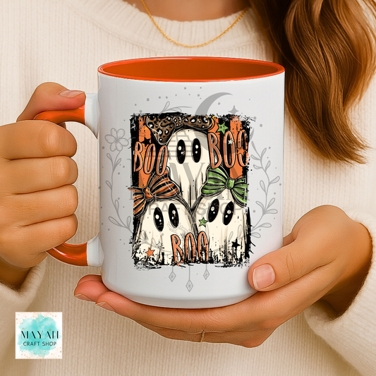 A person holds a white ceramic mug with an orange interior and handle. The mug features a Halloween graphic of four ghosts wearing orange and green bows, with the words 'BOO BOO BOO BOO BAG' in orange.