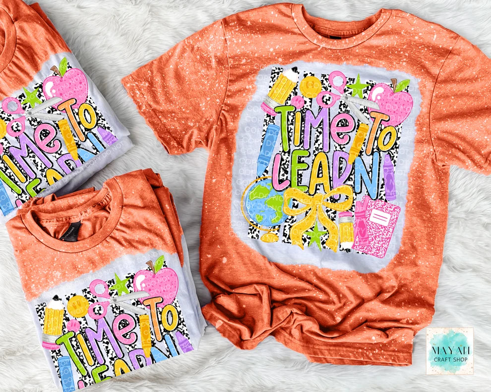 Three coral orange short-sleeve graphic T-shirts arranged on a white faux-fur surface. The shirts feature a colorful school-themed print with the words “Time To Learn,” apples, pencils, a globe, and a notebook.