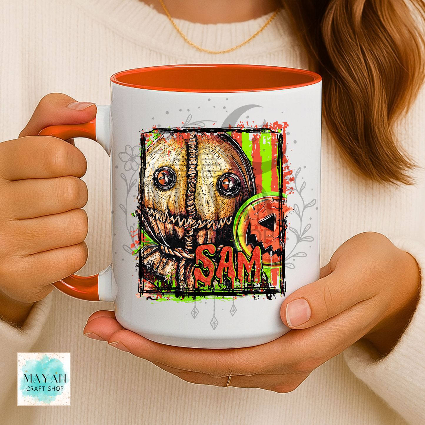 A person holds a white ceramic mug with orange interior and handle, featuring a horror-style illustrated face with a name in orange text and green splatter accents. The person wears a cream knit sweater.