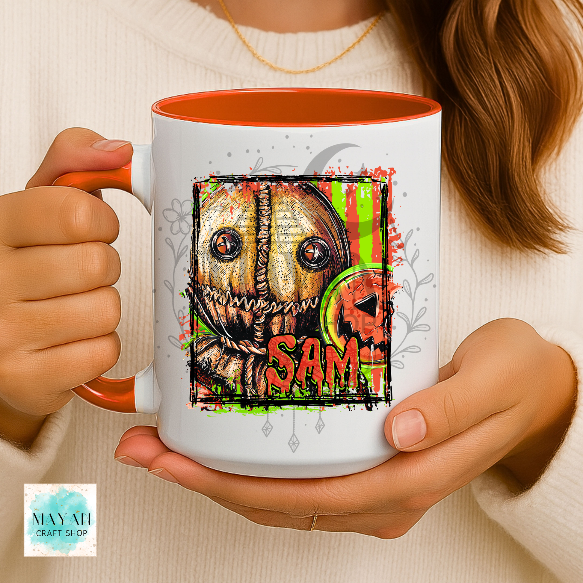 A person holds a white ceramic mug with orange interior and handle, featuring a horror-style illustrated face with a name in orange text and green splatter accents. The person wears a cream knit sweater.