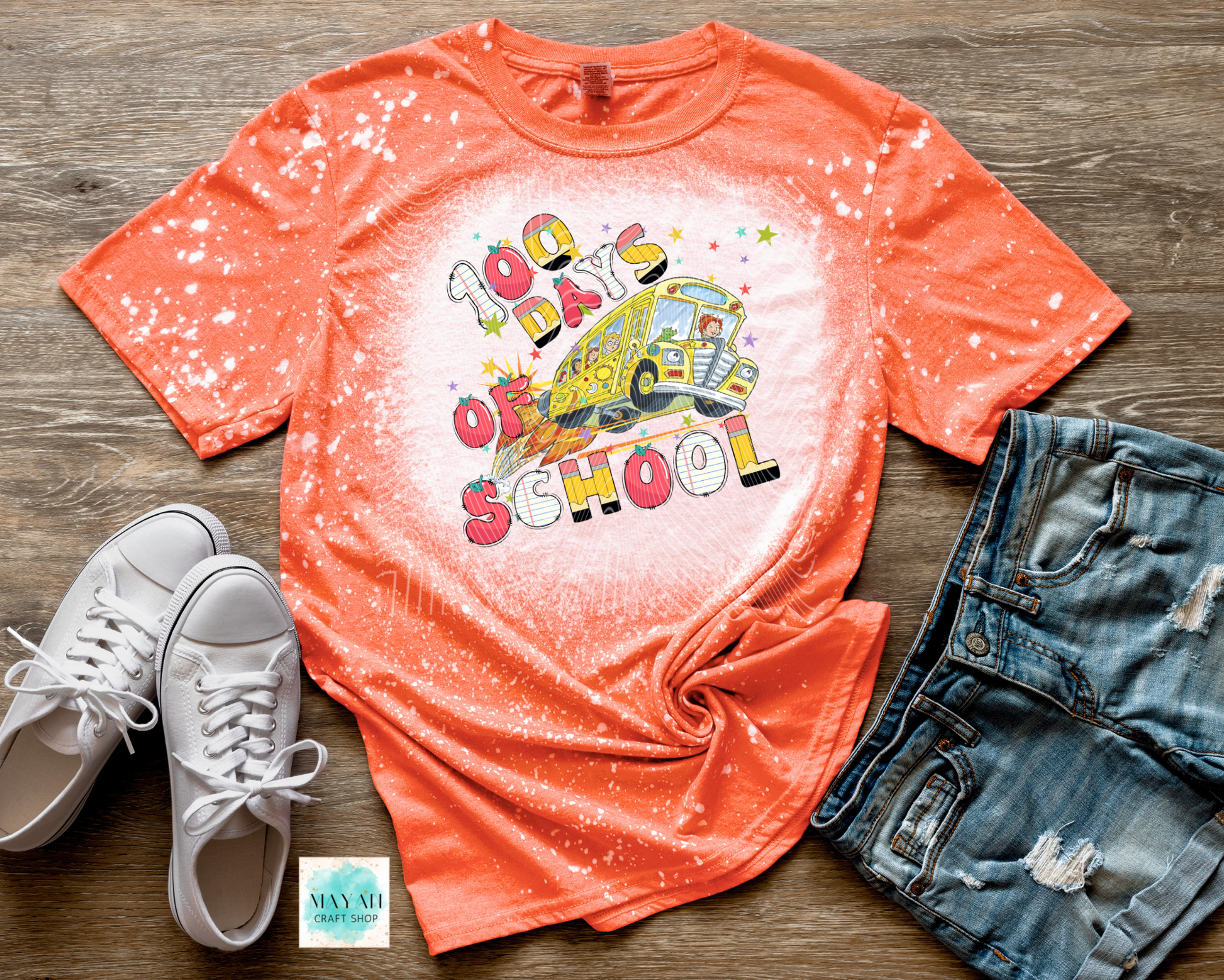 Orange t-shirt with '100 Days of School' graphic, white sneakers, and denim shorts on a wooden surface.