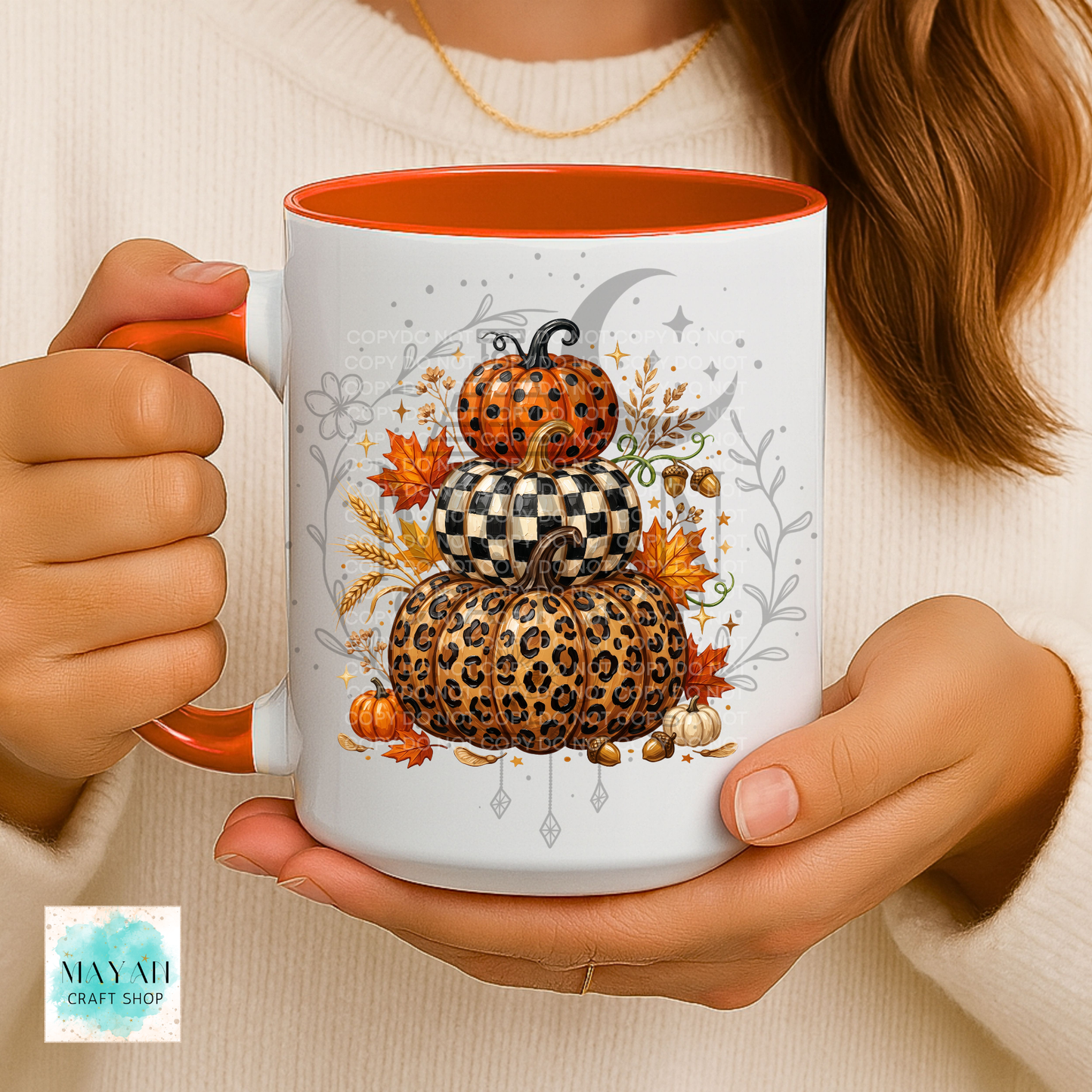 A person holds a white ceramic mug with orange interior and handle, featuring a stacked autumn pumpkin design in leopard print, orange pumpkin, and checkerboard pattern with fall leaves and a crescent moon. The person wears a cream knit sweater.
