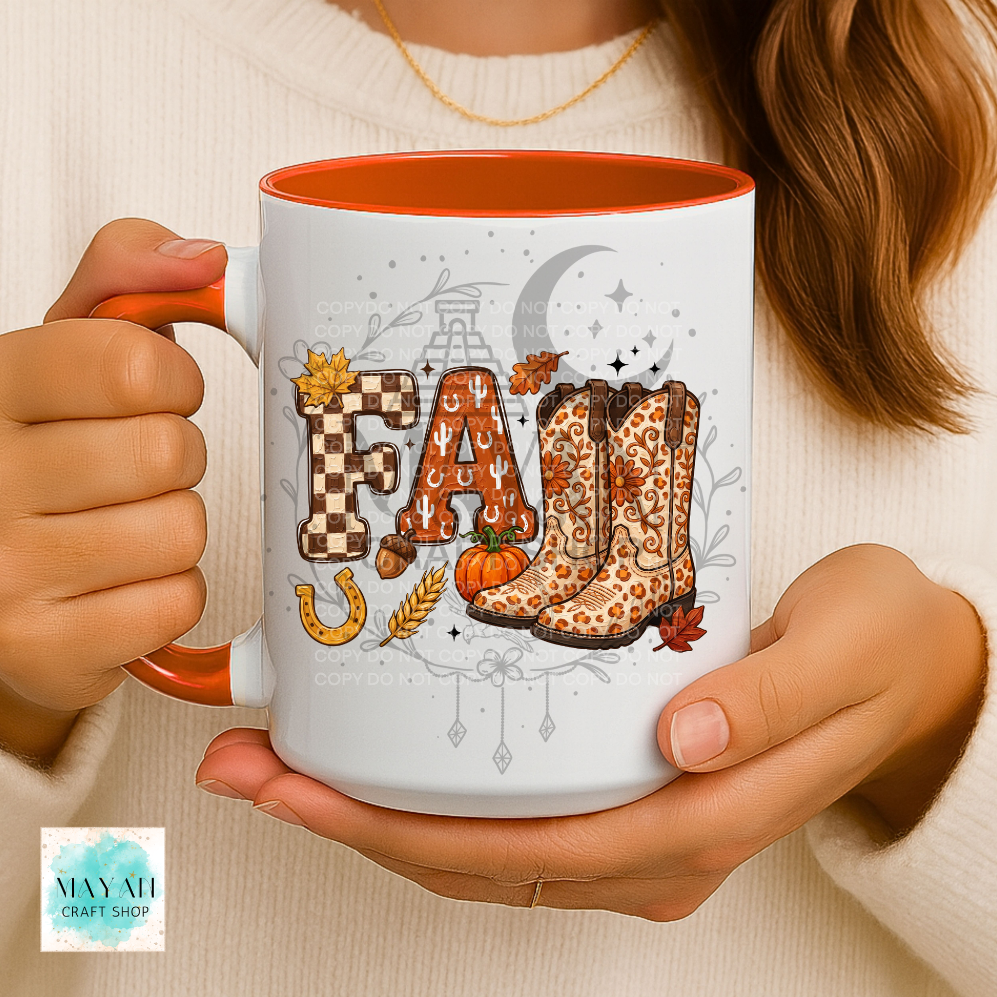 A person holds a white ceramic mug with an orange interior and handle. The mug features a fall-themed graphic spelling 'FALL' with autumn motifs including leopard-print cowboy boots, a pumpkin, wheat, a horseshoe, and a crescent moon.