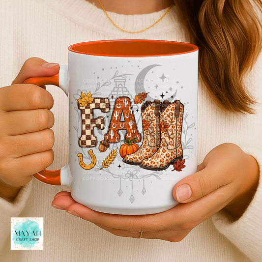A person holds a white ceramic mug with an orange interior and handle. The mug features a fall-themed graphic spelling 'FALL' with autumn motifs including leopard-print cowboy boots, a pumpkin, wheat, a horseshoe, and a crescent moon.