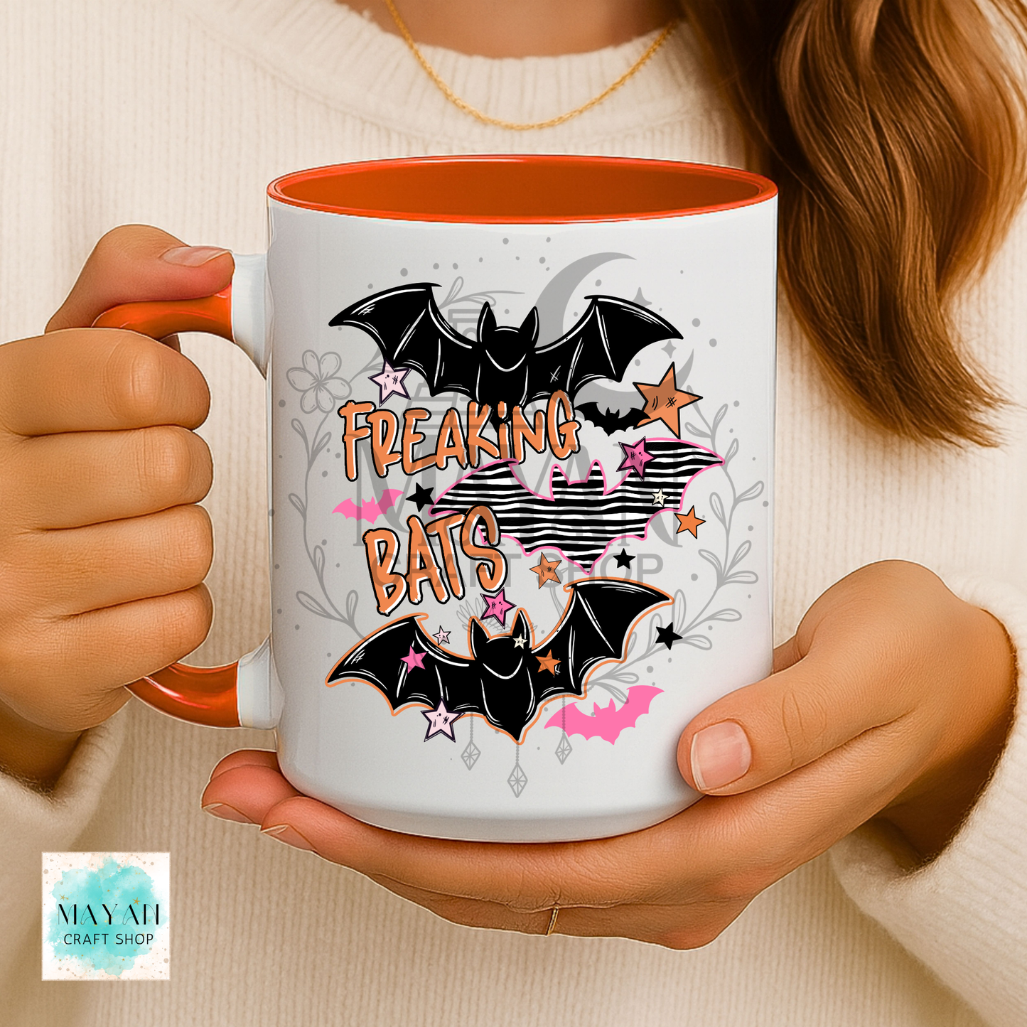 A person holds a white ceramic mug with orange interior and handle. The mug features a Halloween-style graphic with two black bats, the words “FREAKING BATS” in orange, pink stars, gray floral accents, and a cream background.