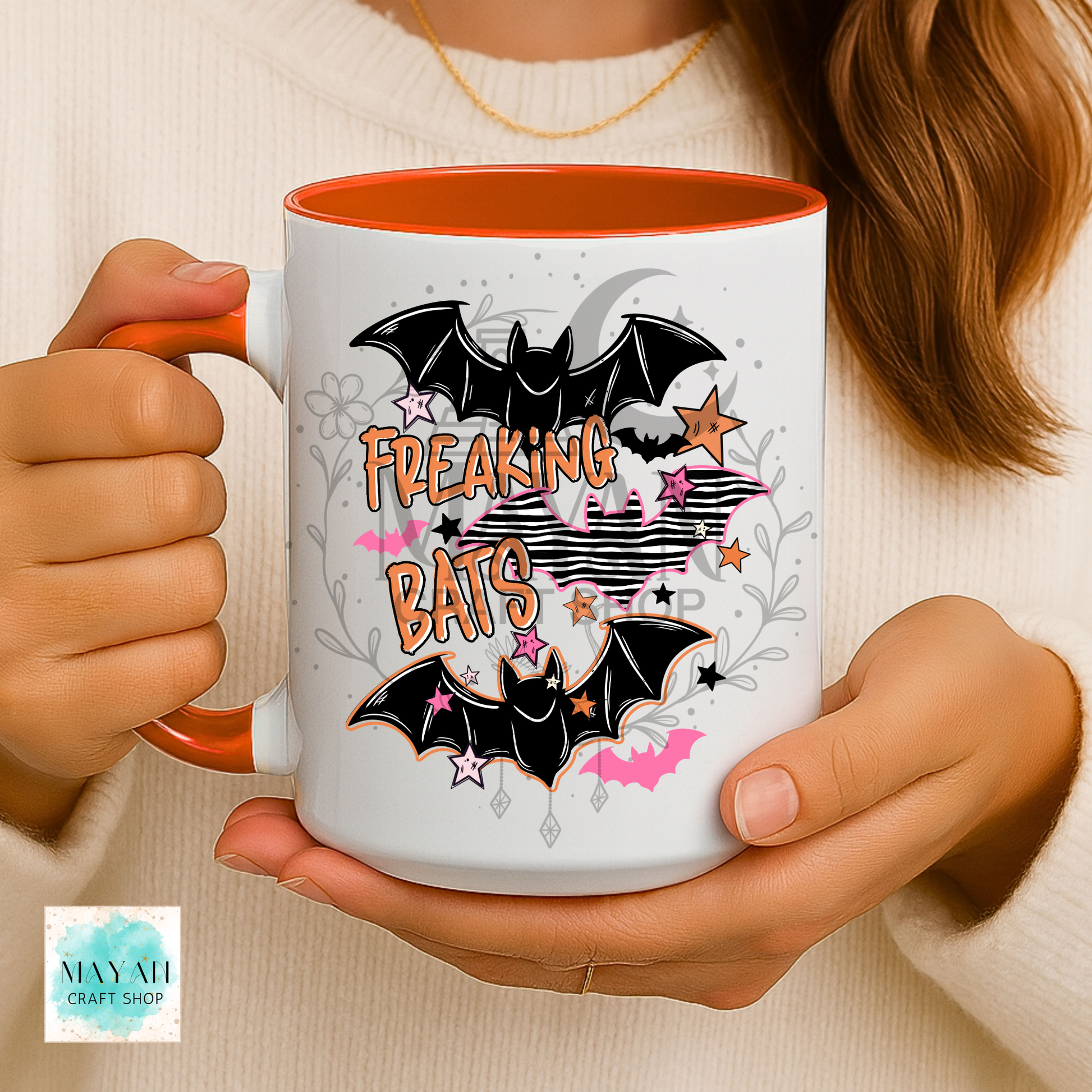 A person holds a white ceramic mug with orange interior and handle. The mug features a Halloween-style graphic with two black bats, the words “FREAKING BATS” in orange, pink stars, gray floral accents, and a cream background.