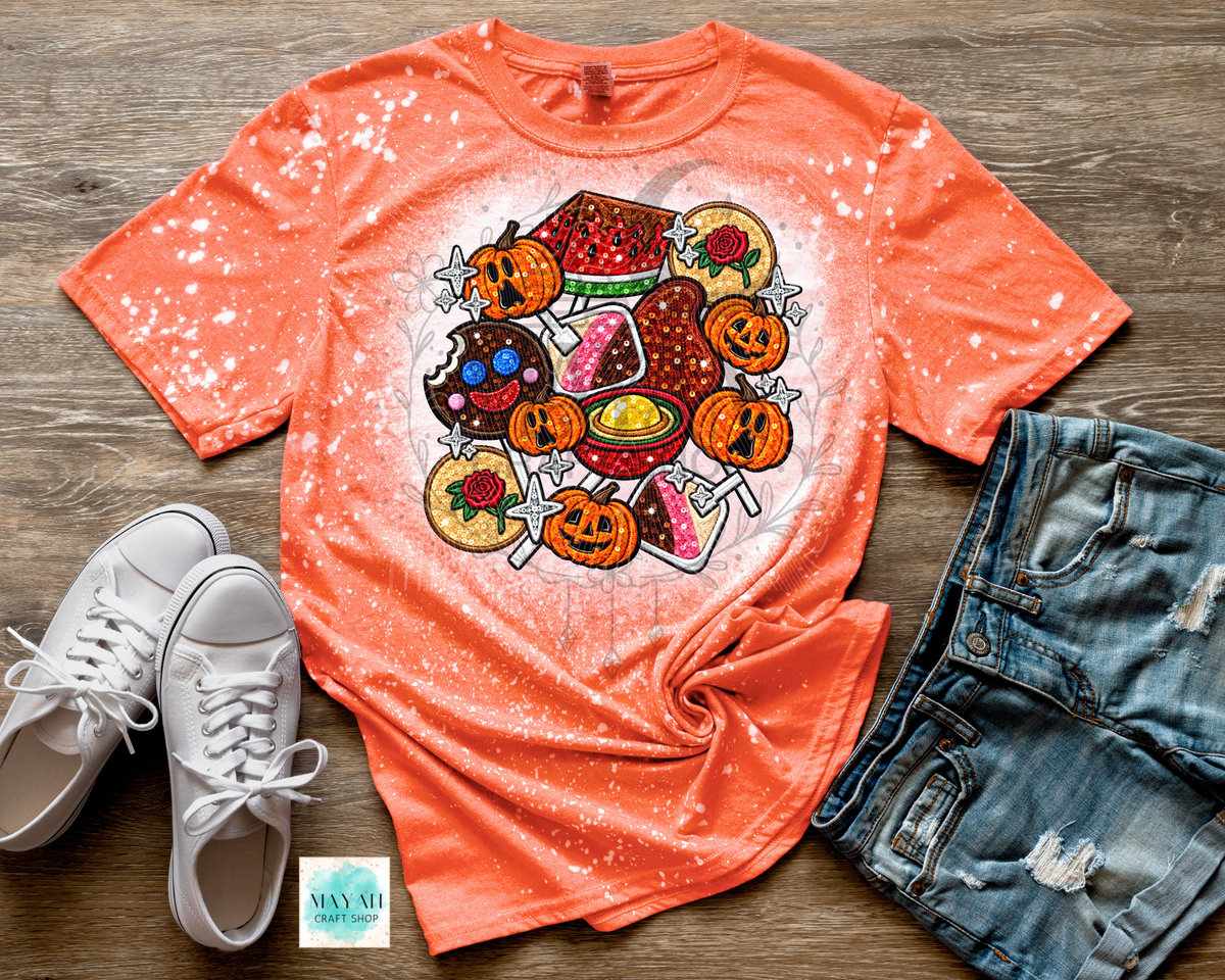 An orange short-sleeve t-shirt with a white bleach-splatter finish and a colorful Halloween-themed graphic print in the center, styled in a flat lay with white sneakers, distressed denim shorts.