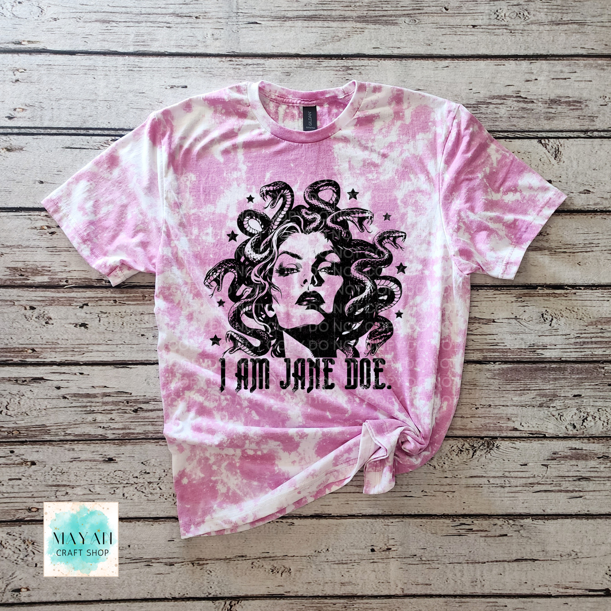 A heather orchid bleached t-shirt featuring a black graphic of Medusa with the text 'I AM JANE DOE.' printed below it, laid flat on a rustic wooden background.