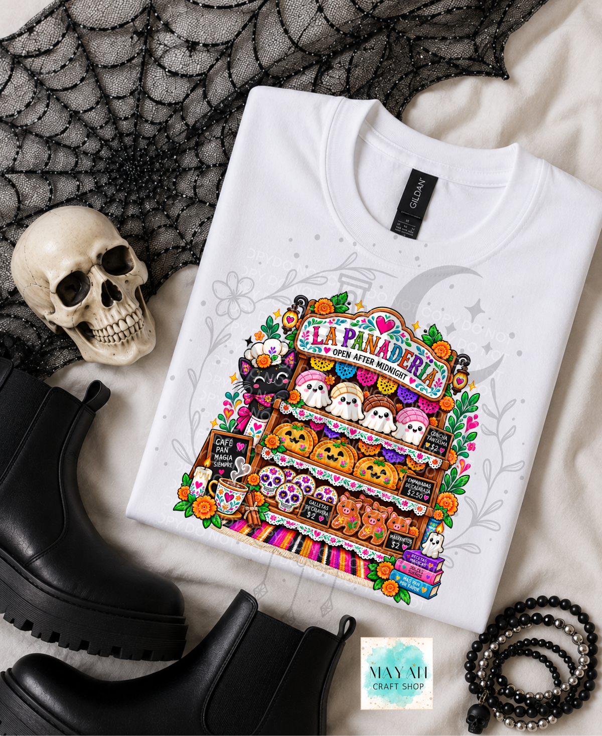 A folded white graphic T-shirt displayed in a styled flat lay with a skull, black boots, spiderweb fabric, and beaded accessories. The shirt features a colorful Halloween-themed illustration of a bakery labeled 'La Panaderia' with ghosts, pumpkins, and treats.