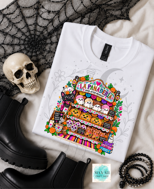 A folded white graphic T-shirt displayed in a styled flat lay with a skull, black boots, spiderweb fabric, and beaded accessories. The shirt features a colorful Halloween-themed illustration of a bakery labeled 'La Panaderia' with ghosts, pumpkins, and treats.
