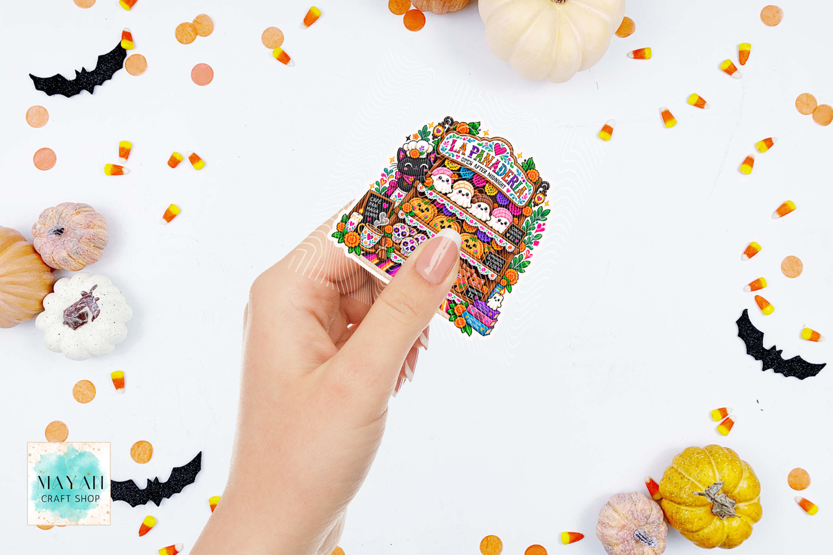 A hand holds a die-cut sticker featuring a colorful Halloween market scene with pumpkins, bats, candy corn, and cartoon ghosts. The background is white with scattered Halloween decorations, including pumpkins, bats, and candy corn.