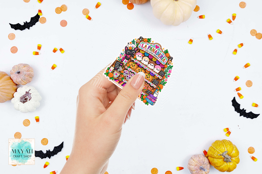 A hand holds a die-cut sticker featuring a colorful Halloween market scene with pumpkins, bats, candy corn, and cartoon ghosts. The background is white with scattered Halloween decorations, including pumpkins, bats, and candy corn.