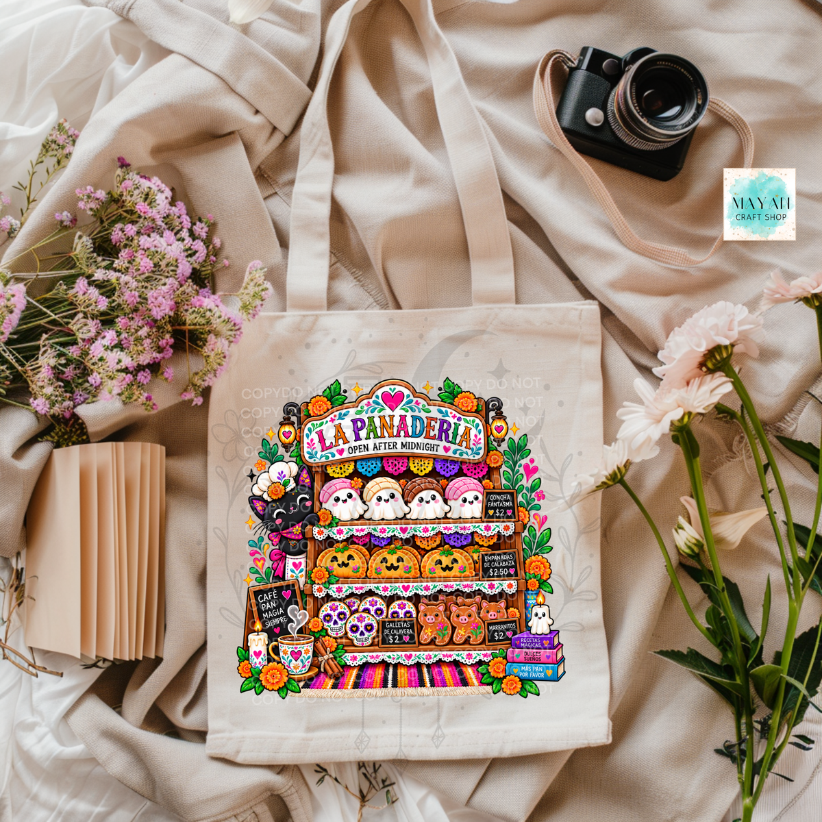 A beige canvas tote bag with a colorful 'La Panadería' cartoon bakery illustration, styled in a flat lay with a vintage camera, pink flowers, and cream fabric.