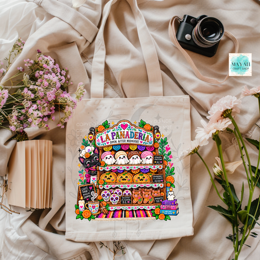 A beige canvas tote bag with a colorful 'La Panadería' cartoon bakery illustration, styled in a flat lay with a vintage camera, pink flowers, and cream fabric.