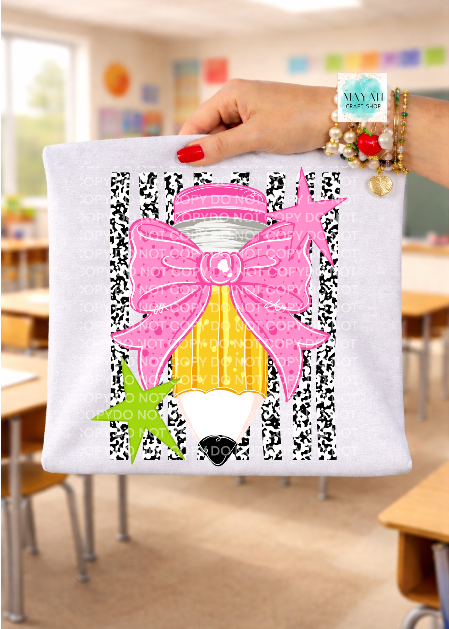 A hand holds up a white T-shirt featuring a colorful pencil graphic with a pink bow, yellow striped body, black-and-white patterned background, and decorative text. The background shows a blurred classroom setting.
