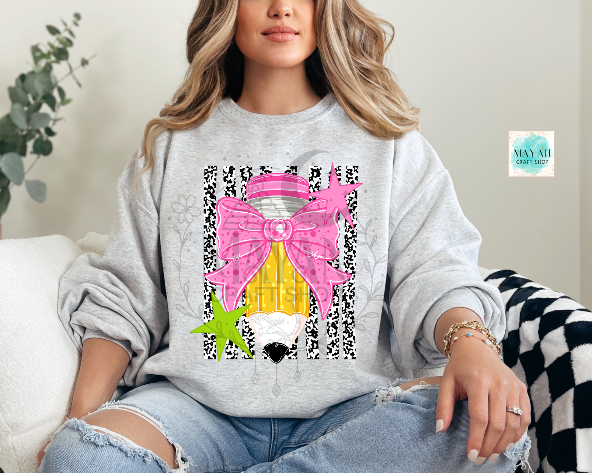 A woman wearing a light gray sweatshirt with a colorful graphic of a pencil, pink bow, and green starfish, paired with ripped blue jeans, sitting on a couch.