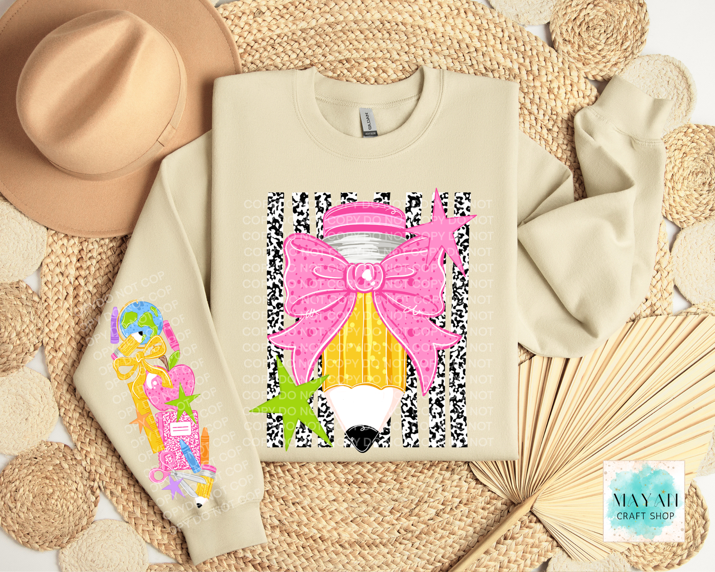 A folded sand sweatshirt with colorful graphic prints, styled in a flat lay with a tan hat, woven mat, and paper fan. The sweatshirt features a large pink bow and yellow striped pencil design on the front, and a pastel multicolor design on the left sleeve.
