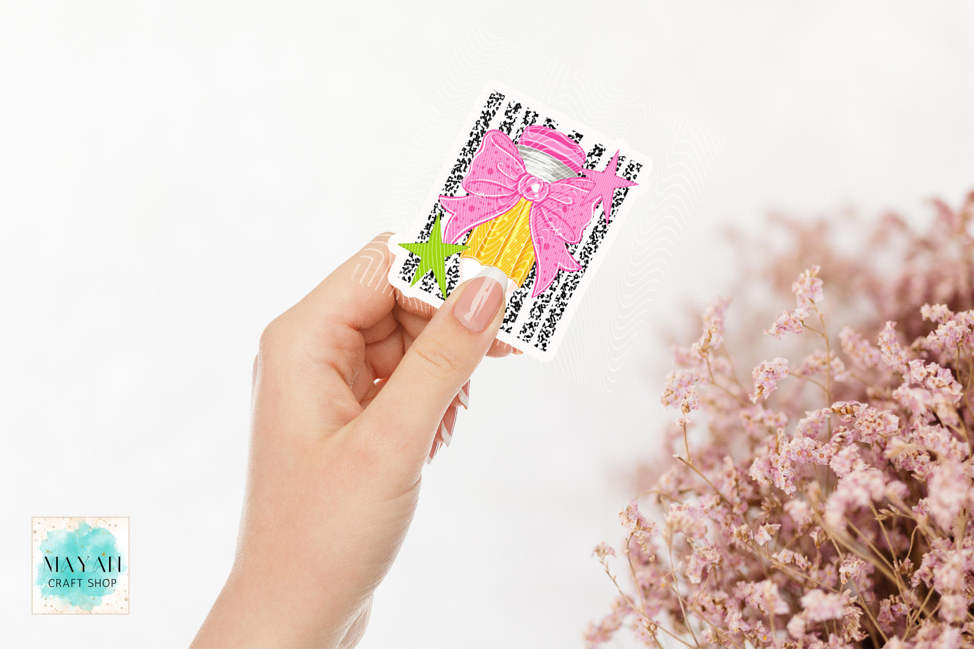 A hand holds a small square craft sticker featuring a pink bow, yellow dress, and green star on a black patterned background. The scene is set against a clean white background with soft pink dried flowers.