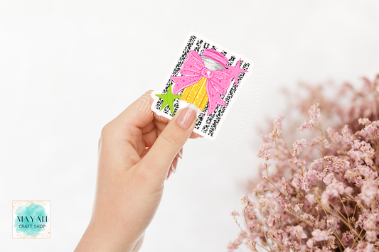 A hand holds a small square craft sticker featuring a pink bow, yellow dress, and green star on a black patterned background. The scene is set against a clean white background with soft pink dried flowers.