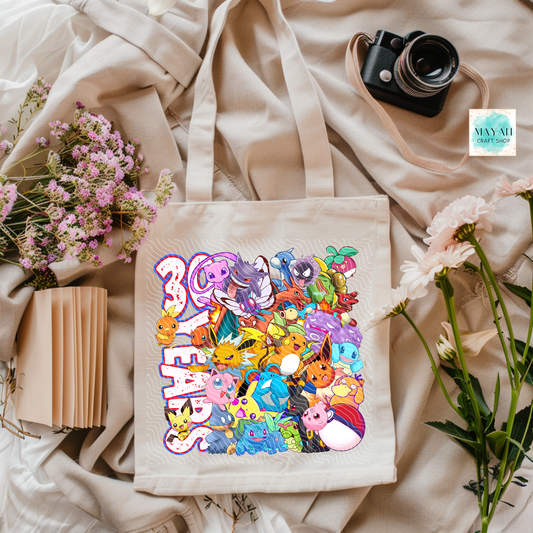 Tote bag with colorful character design and  text, surrounded by flowers and a camera on a light fabric background.