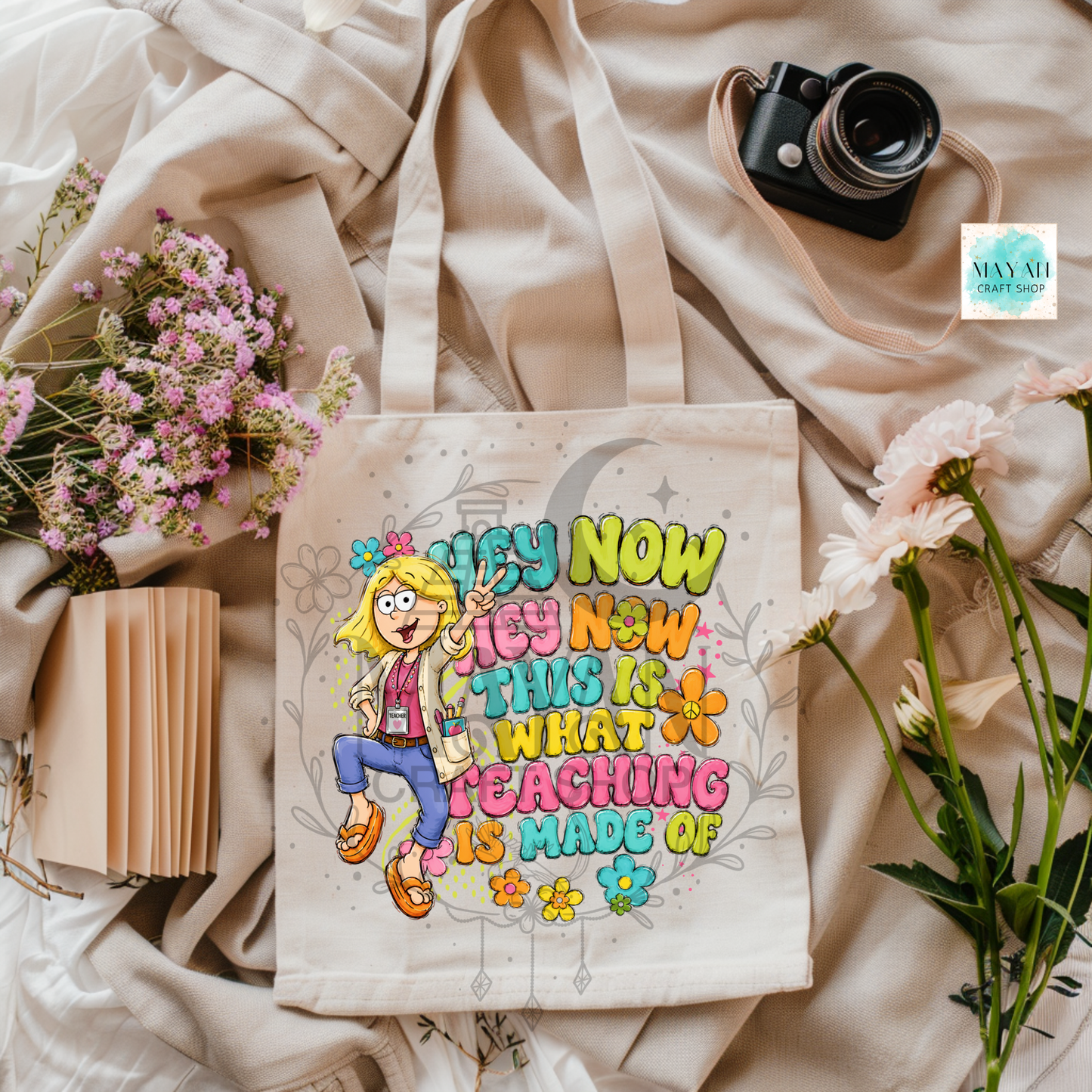 A cream canvas tote bag with a colorful cartoon girl and the text 'TEACHING THIS IS WHAT I DO NOW' is styled in a flat lay with pink flowers, a book, a camera, and draped beige fabric. 