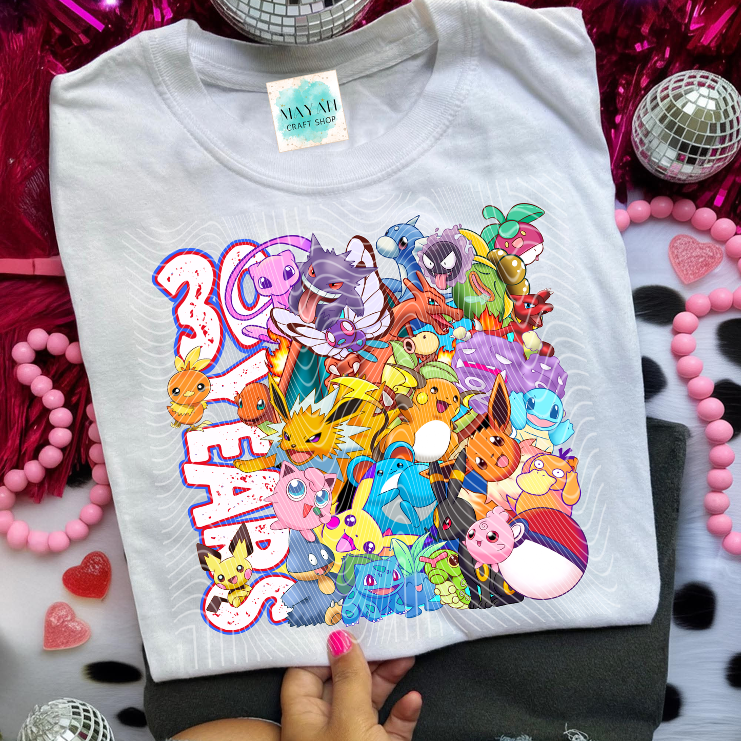 White t-shirt with colorful cartoon characters and text held by a hand against a festive background.