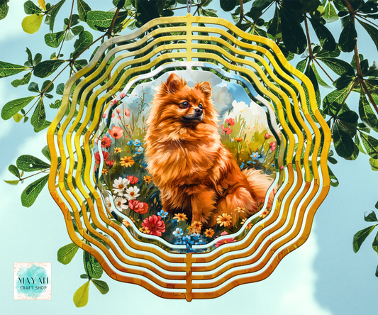 A wind spinner, featuring a printed illustration of a fluffy orange dog in a flower meadow. The piece is displayed against a background of green leaves and a light blue sky.
