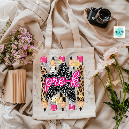 A canvas tote bag with a graphic design of four pencils in leopard print and pink caps, featuring the text 'pre-k' in pink script over a black star. The bag is styled in a flat lay with beige fabric, flowers, a camera, and a notebook.