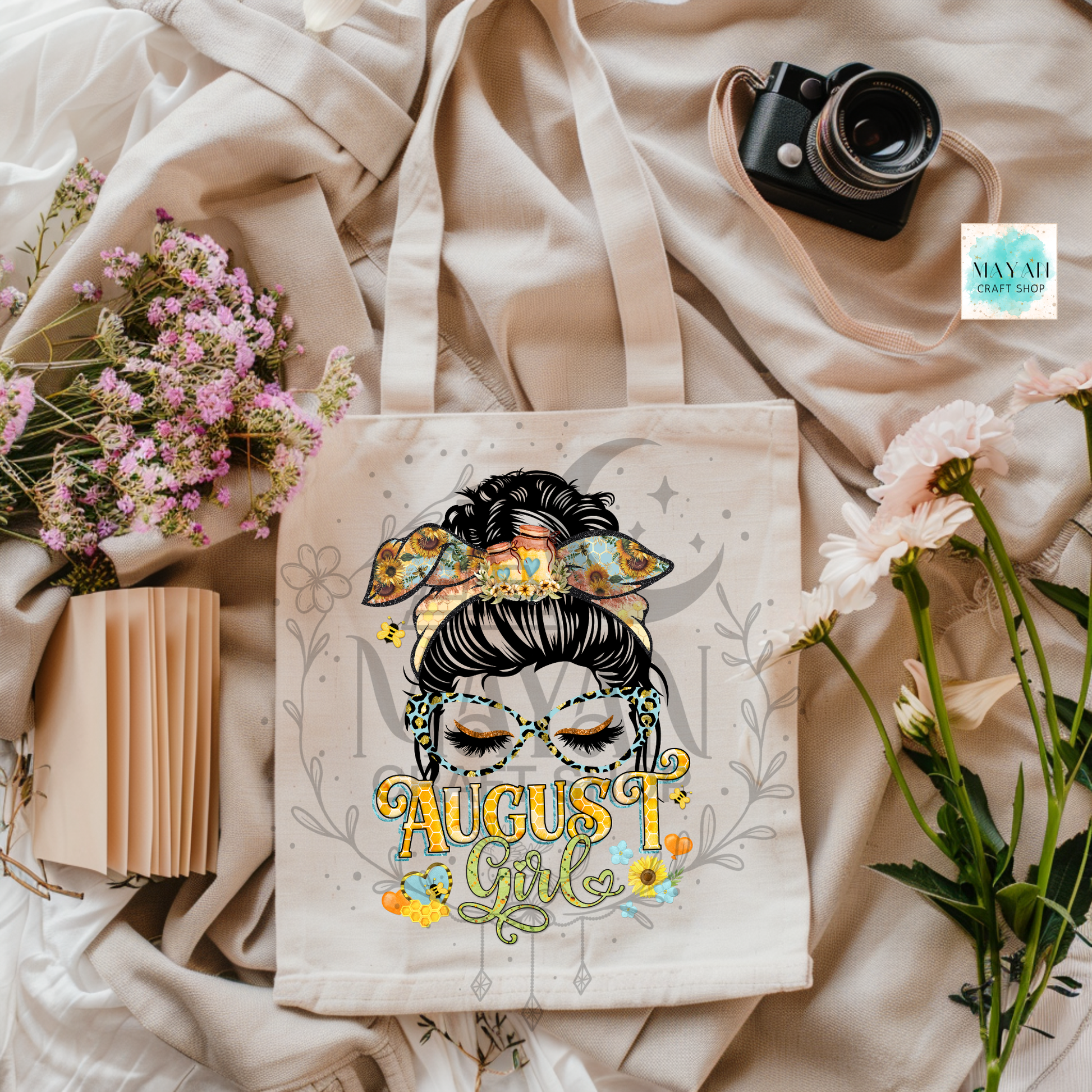 A cream canvas tote bag with a colorful graphic of a woman in glasses and the text 'AUGUST Girl' surrounded by flowers, styled in a flat lay with a book, flowers, a camera, and draped fabric.