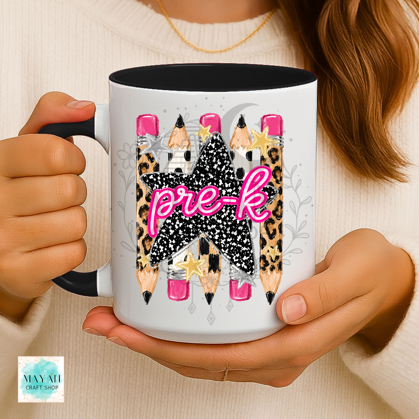 A person holds a white ceramic mug with a black handle and interior. The mug features a graphic design of pencils with leopard print, black glitter textures, pink tips, and the word 'pre-k' in bright pink script.