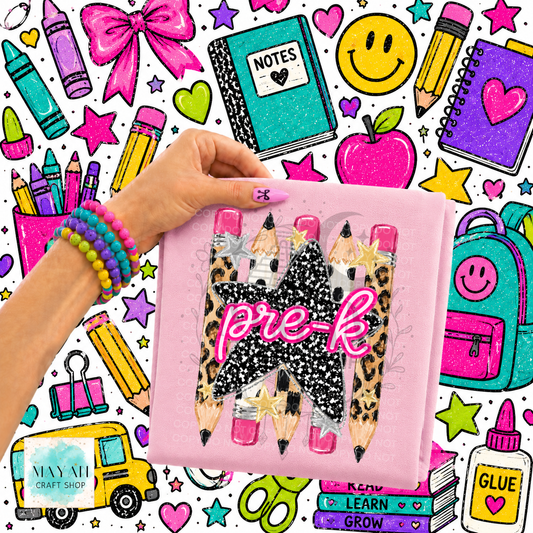 A hand holds a pink shirt featuring three decorative pencils with leopard print, black glitter, and pink glitter tips, with the words 'pre-k' in pink glitter. The background is a busy, colorful school-supplies illustration with crayons, notebooks, an apple, stars, and a yellow bus.