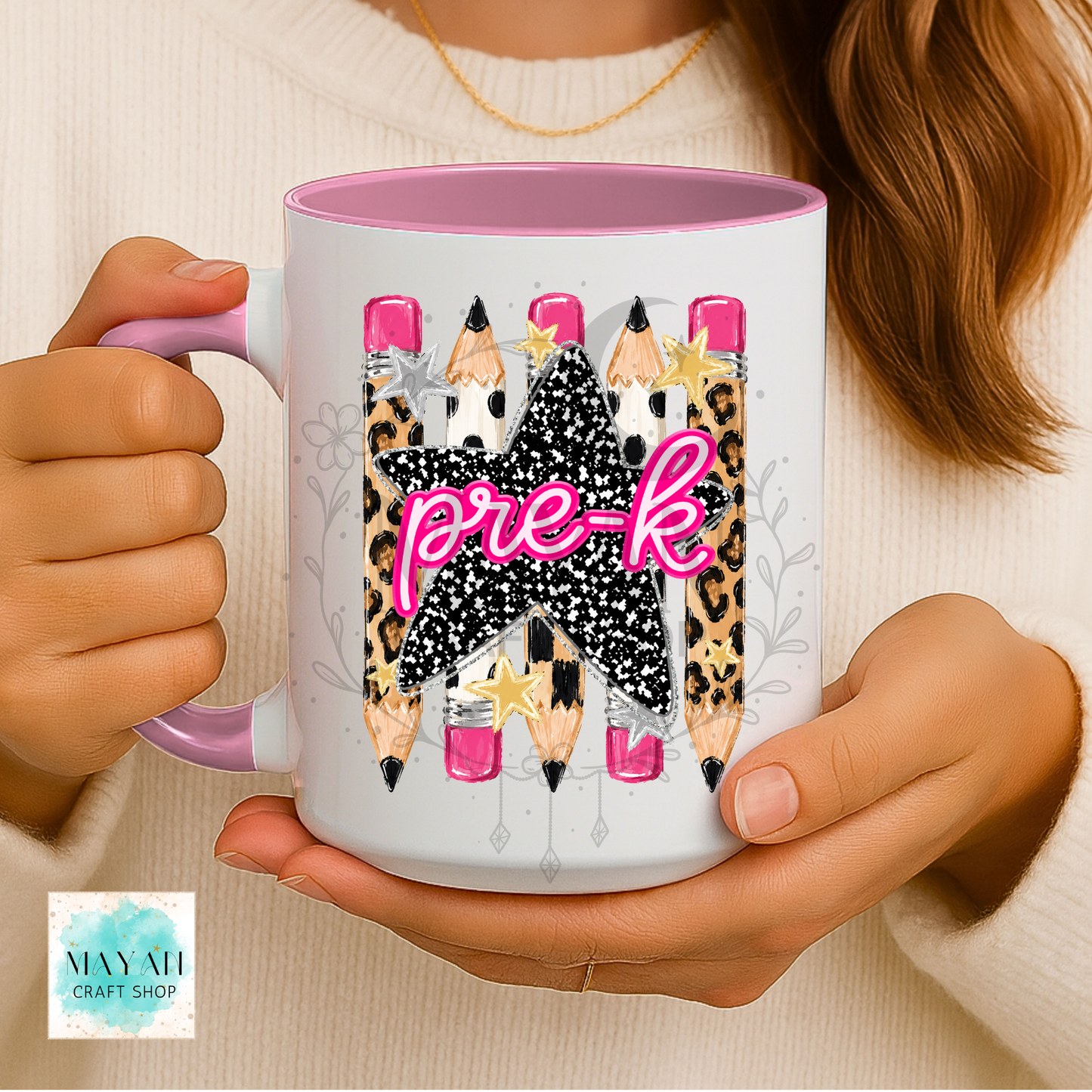A person holds a white ceramic mug with a pink interior and handle. The mug features a graphic of three pencils with leopard print, black-and-white speckled patterns, pink tips, and the word 'pre-k' in pink script.