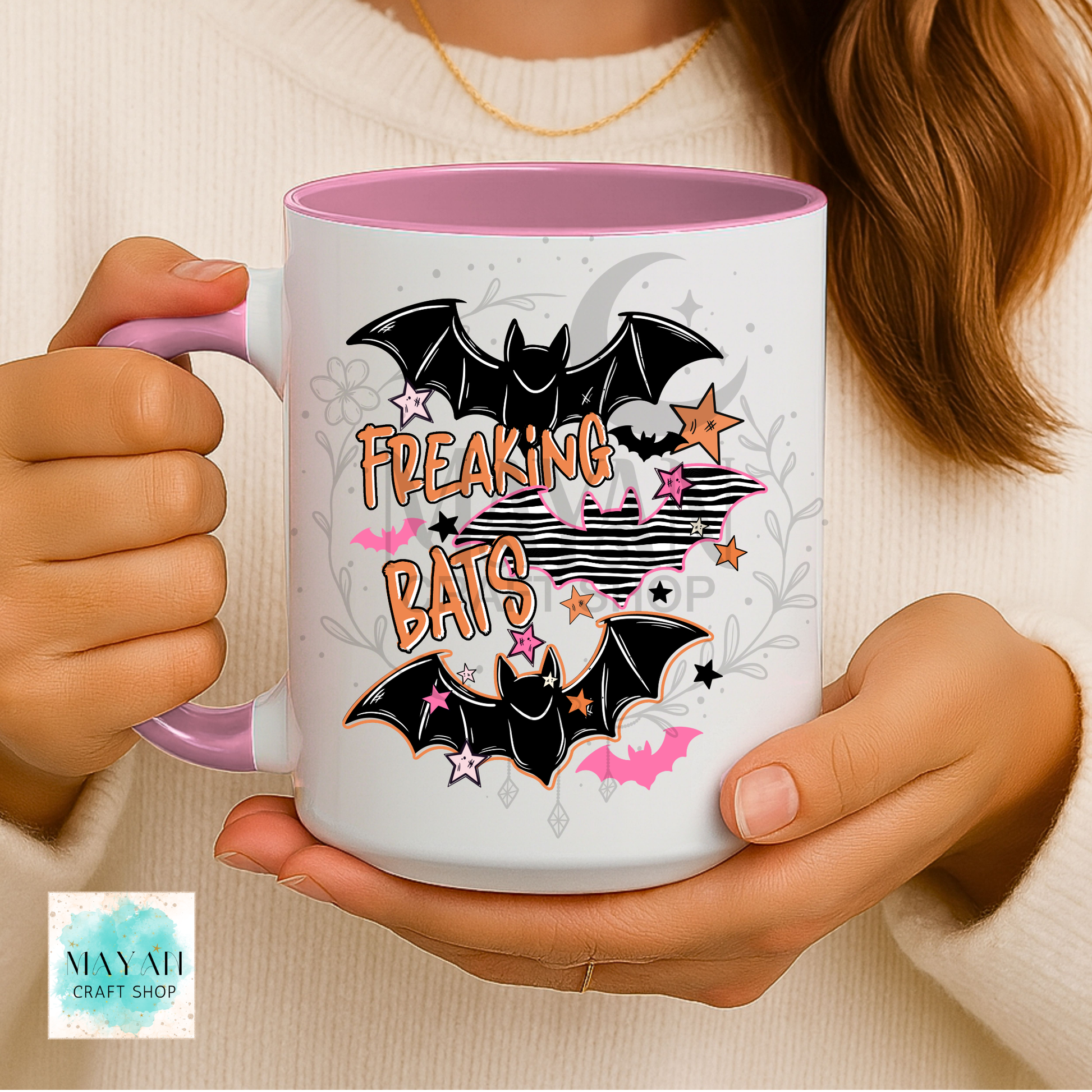 A person holds a white ceramic mug with a pink interior and handle. The mug features a Halloween-themed graphic with two black bats, pink stars, and the words 'FREAKING BATS' in orange.