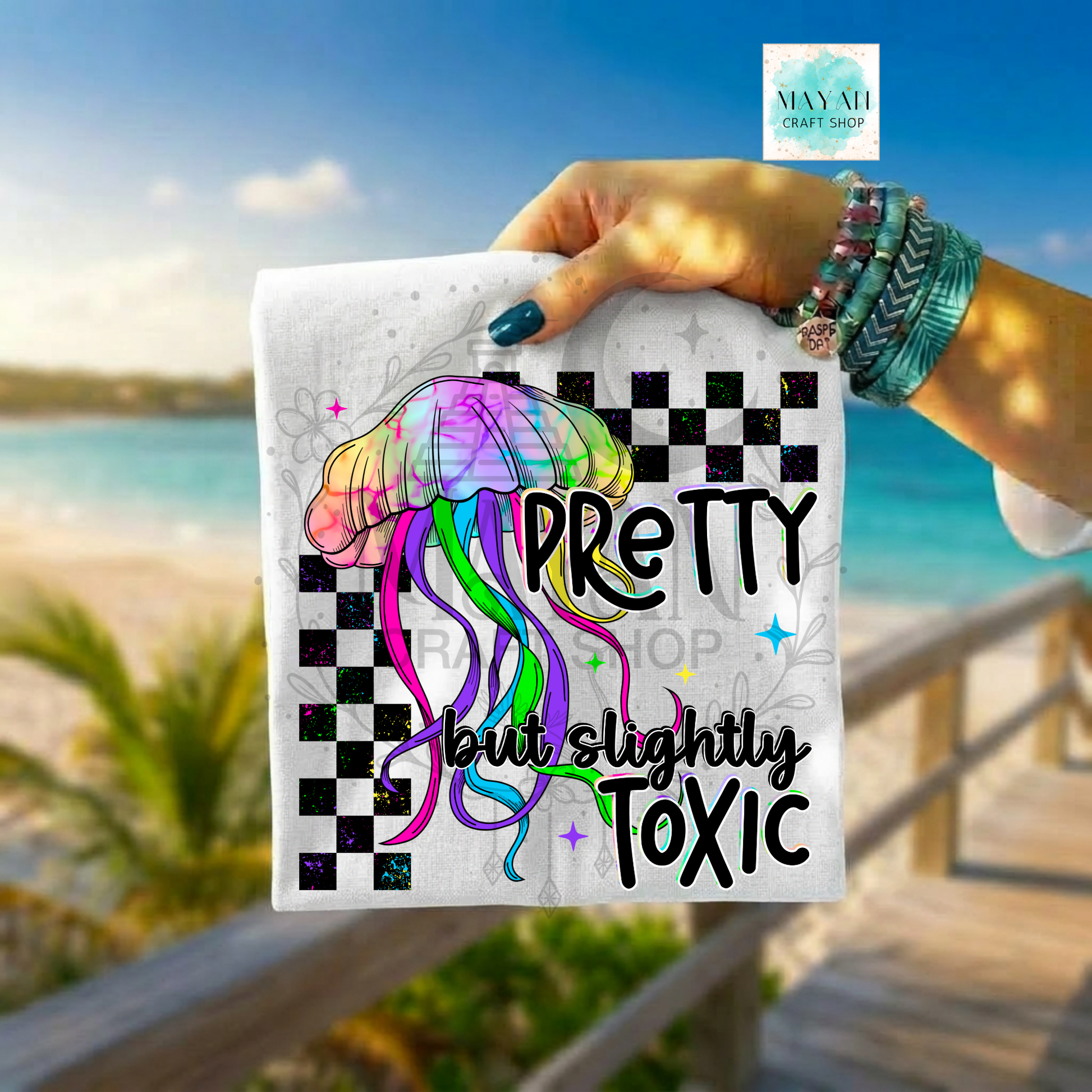 Hand holding a colorful jellyfish shirt with text by a beach