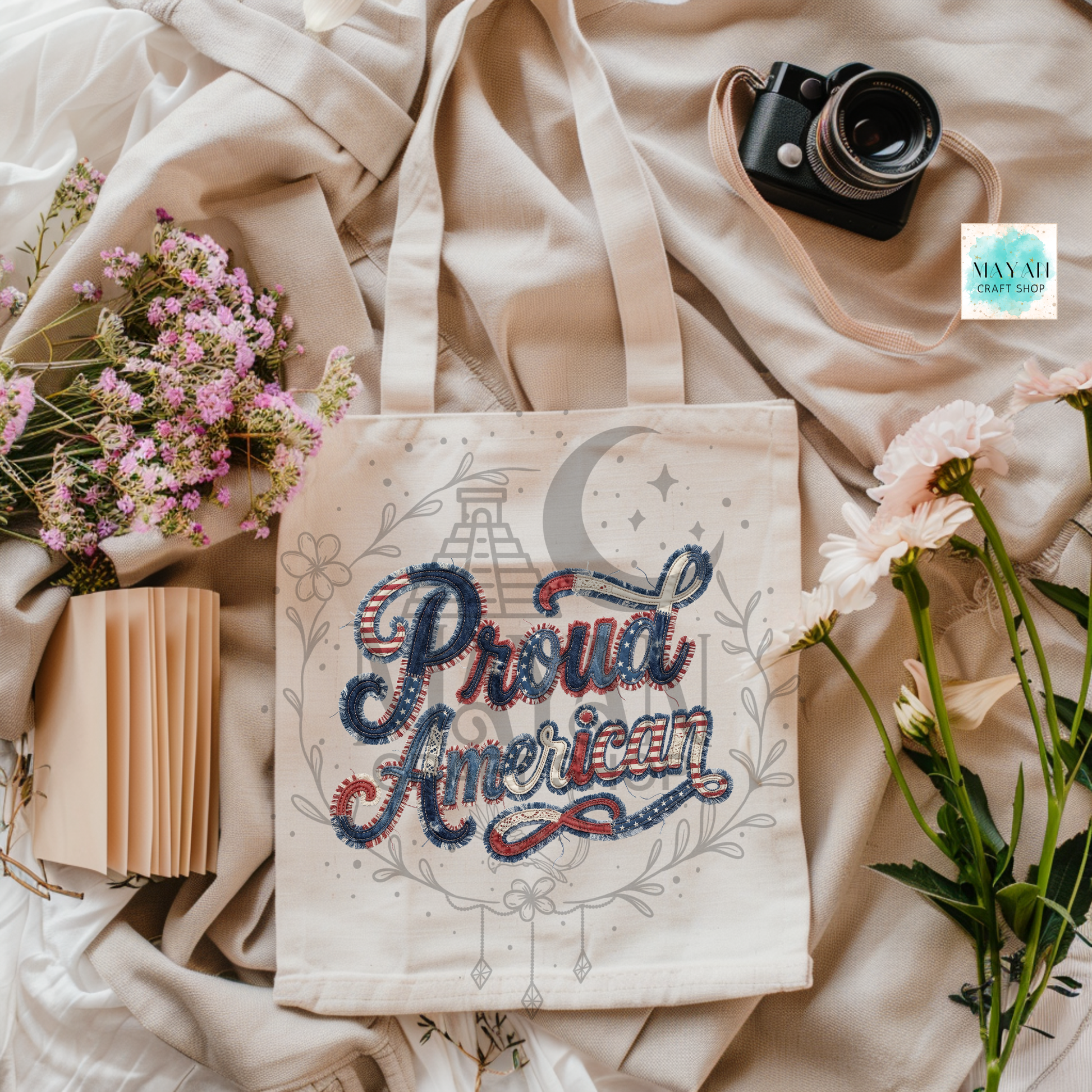 Tote bag with 'Proud American' design on a soft fabric background with flowers and a camera.