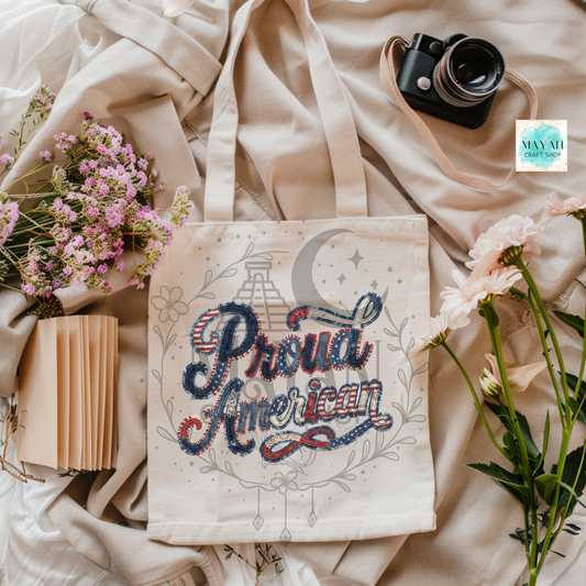 Tote bag with 'Proud American' design on a soft fabric background with flowers and a camera.