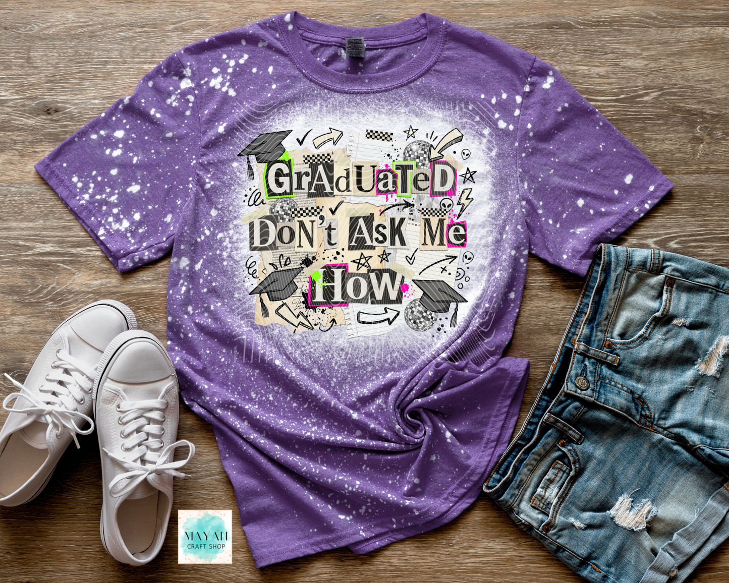 Purple t-shirt with graduation design on a wooden surface with shoes and jeans.