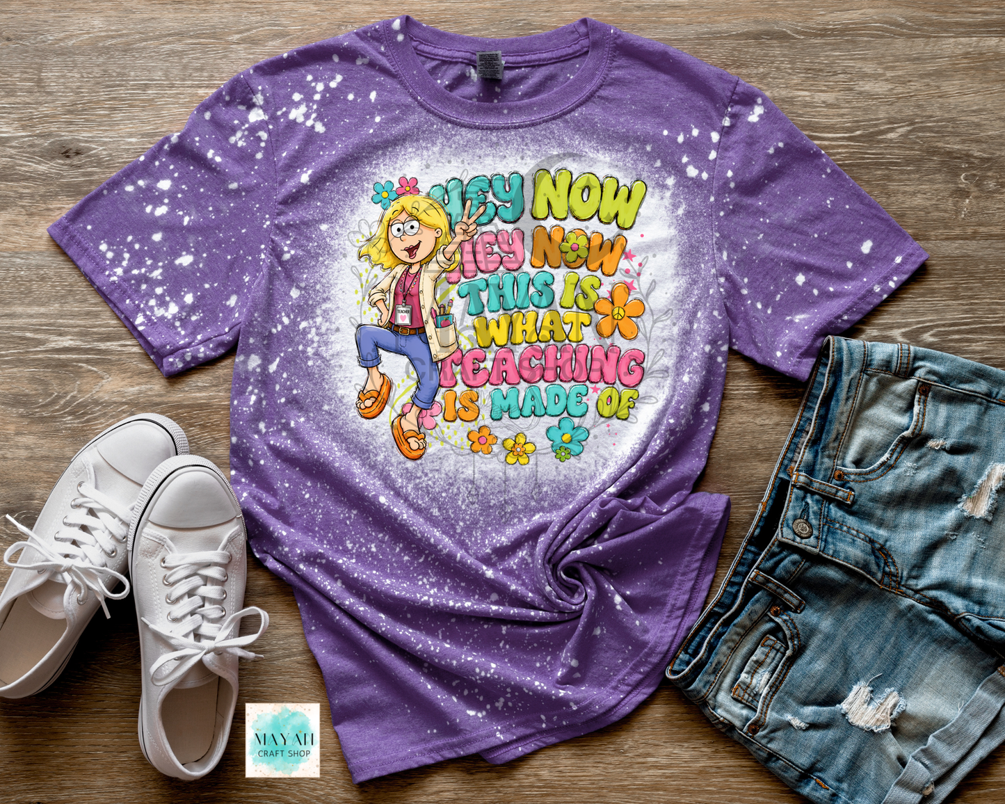 A purple short-sleeve graphic T-shirt with a white bleach-splatter finish and a colorful cartoon girl design, styled in a flat lay on a wood surface with white sneakers, distressed denim shorts.