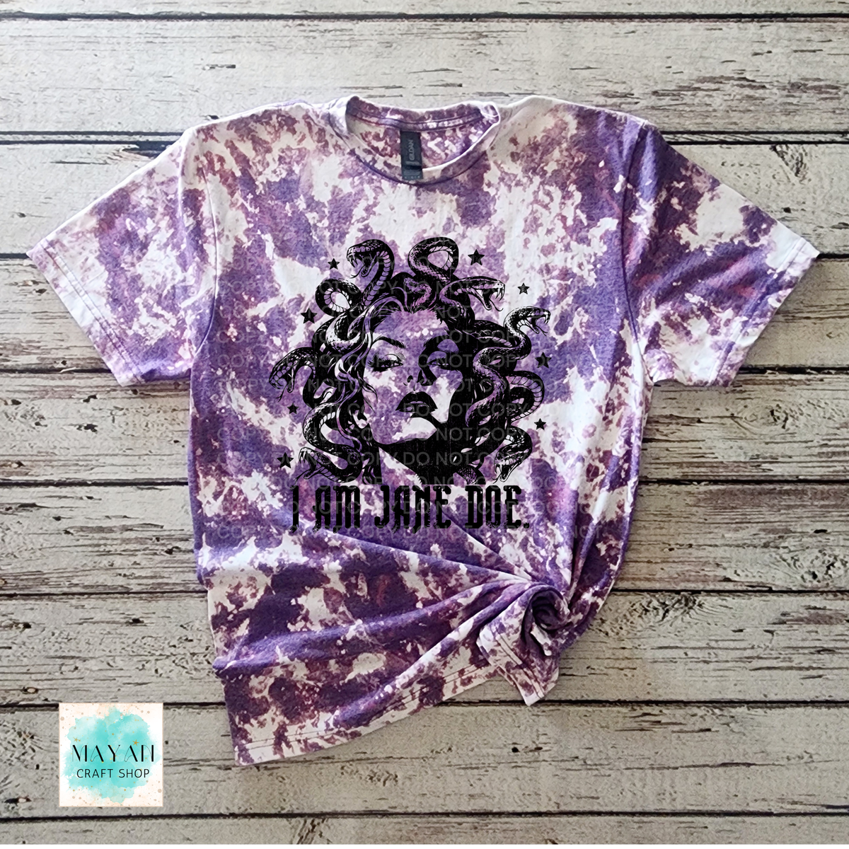 A heather purple bleached t-shirt with a black graphic of Medusa's face and the text 'I AM JADE DOE' printed on the front, displayed flat on a rustic wooden background.