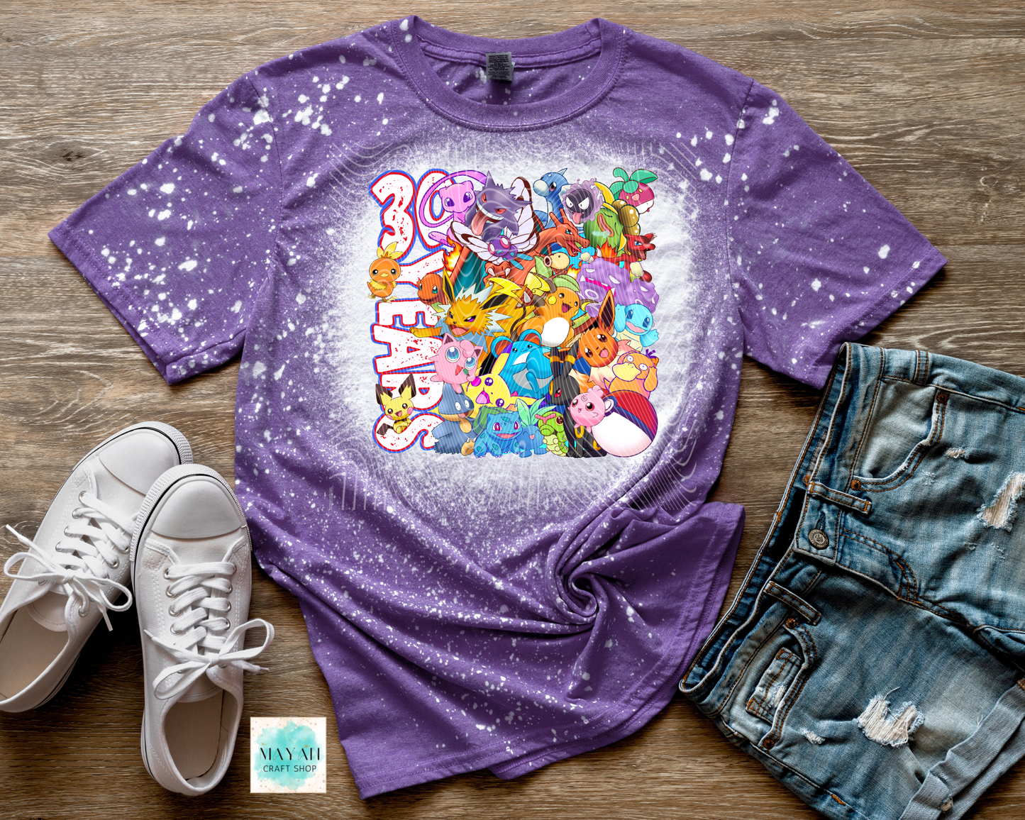 Purple t-shirt with colorful graphic design on a wooden surface with shoes and shorts.