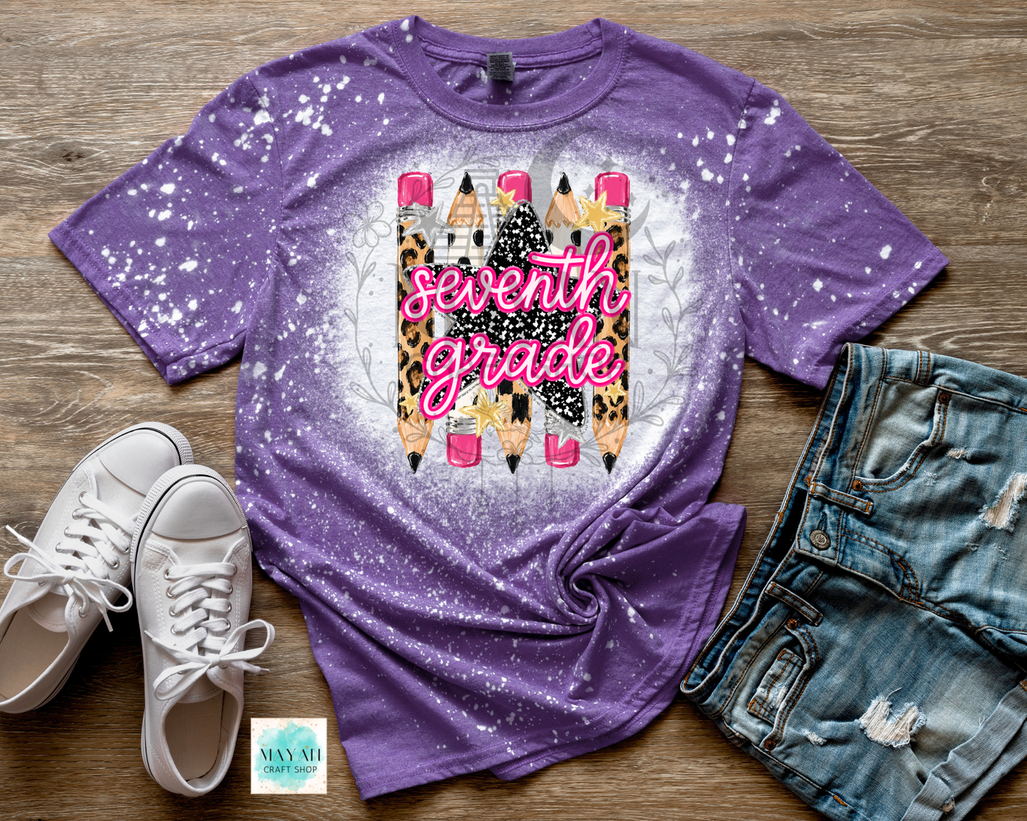 A purple short-sleeve t-shirt with a white bleach-splatter finish and a central graphic of colorful pencils and the words 'seventh grade' in pink script. The shirt is styled in a flat lay on a wooden surface alongside white sneakers, denim shorts.