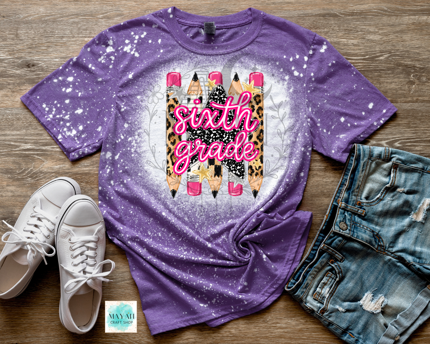 A purple short-sleeve t-shirt with a white bleach-splatter finish and a 'sixth grade' graphic featuring colorful pencils, styled in a flat lay with white sneakers, denim shorts.