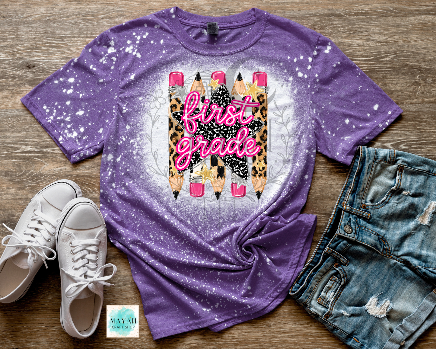 A purple short-sleeve graphic T-shirt laid flat on a wood surface, styled with white sneakers, distressed denim shorts. The shirt has a white splatter/dye effect and a central graphic featuring pink script text reading 'first grade' over leopard-print pencils and a white oval background.