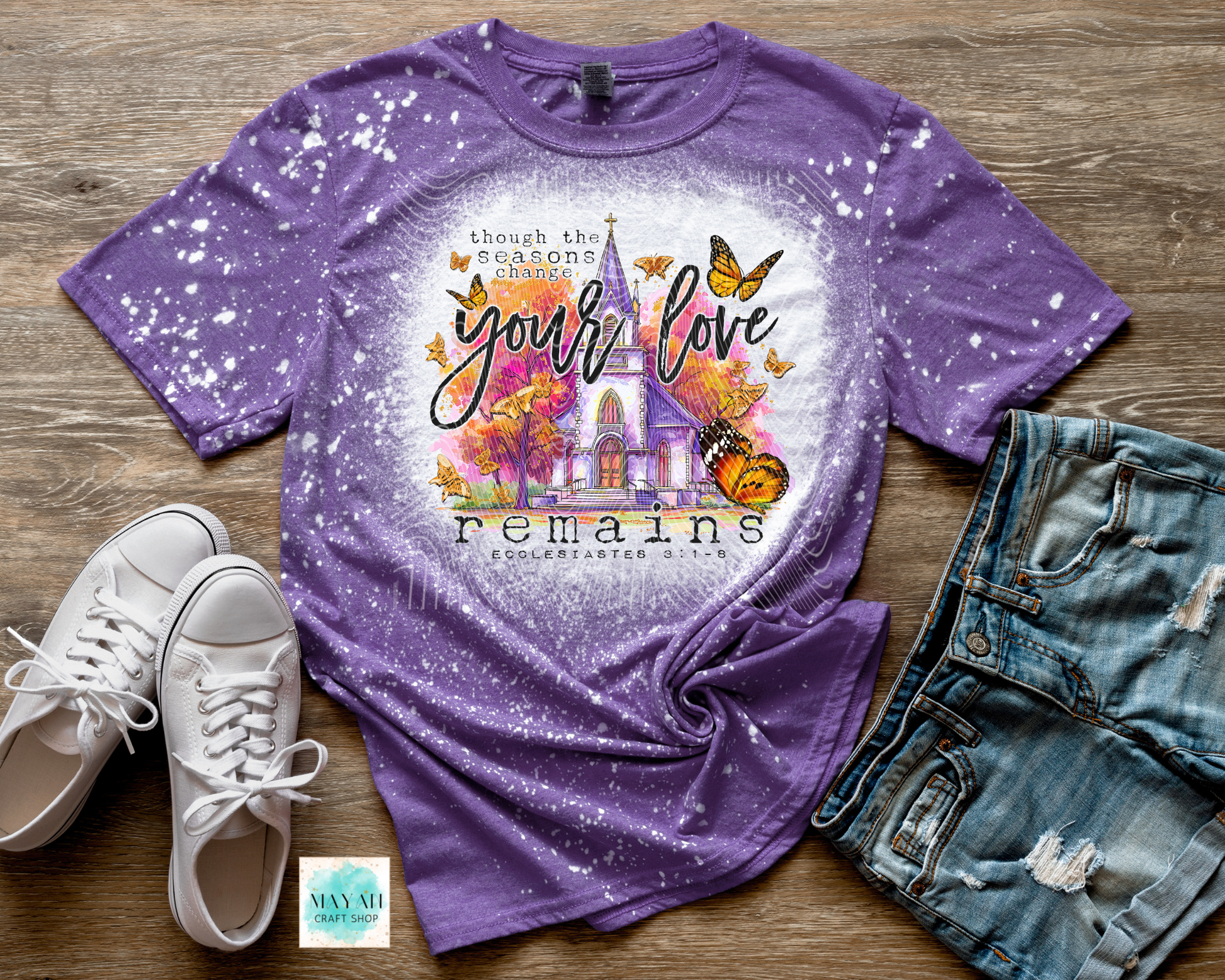 Purple t-shirt with a colorful design and text on a wooden surface with shoes and jeans.