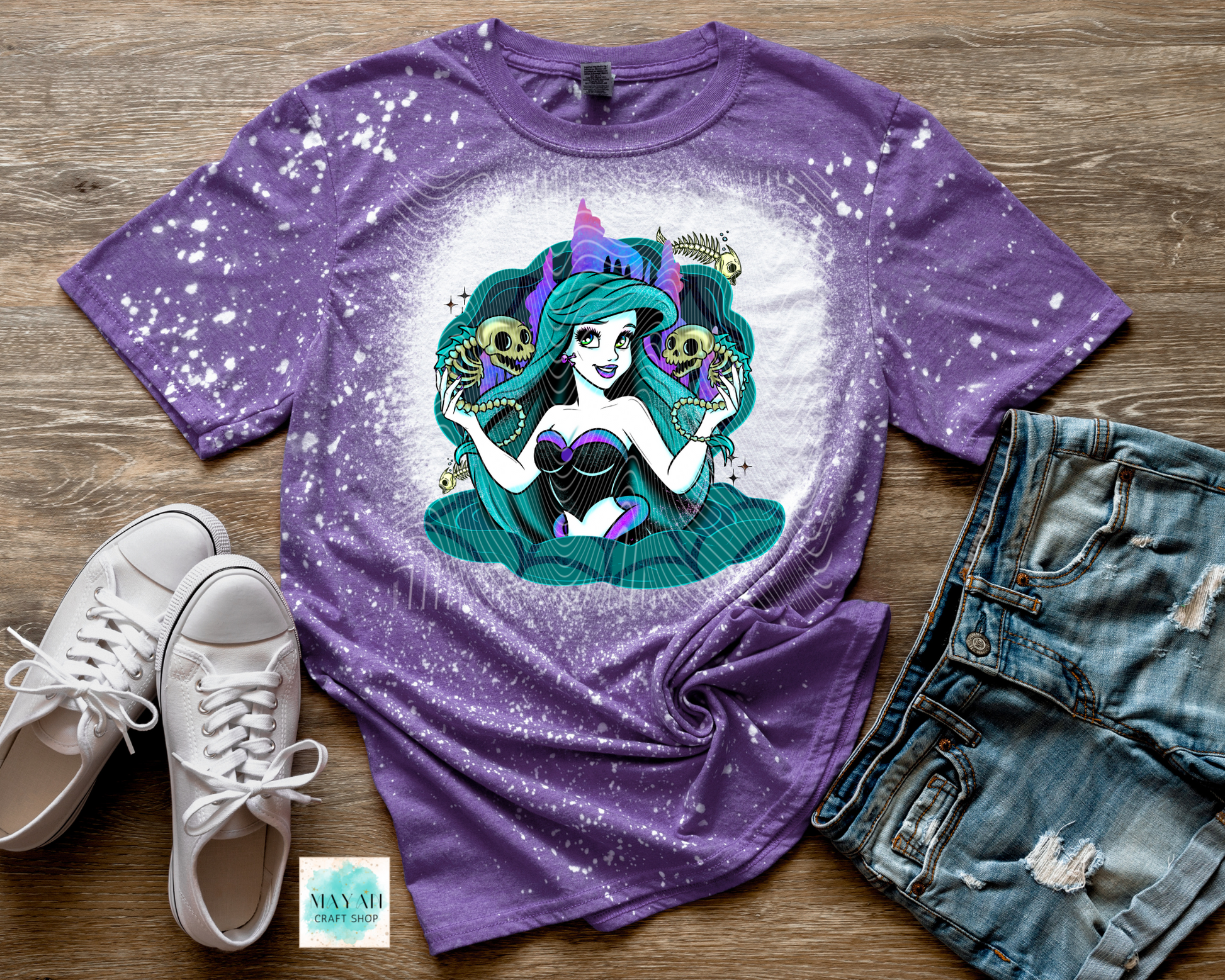 Purple t-shirt with a mermaid graphic on a wooden surface with sneakers and shorts.